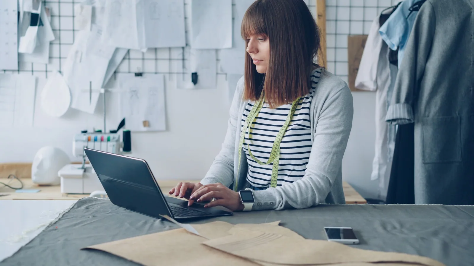 Fashion designer working on a laptop beside sketches in a bright studio